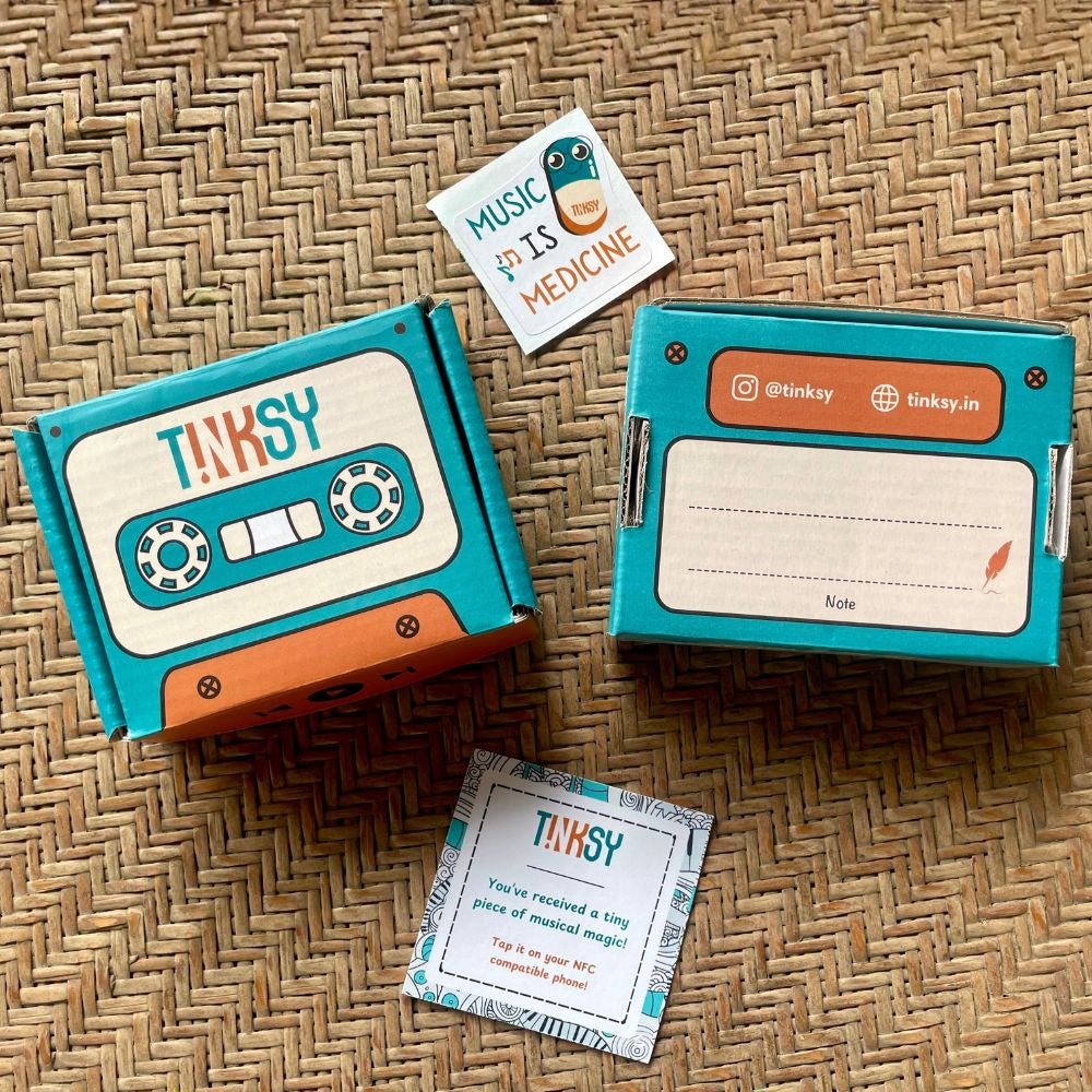 The image features two colorful boxes from TINKSY, designed with a retro cassette tape theme. One box has a label for a note, while a small card nearby reads Youve received a tiny piece of musical magic! and encourages tapping for music.